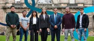 Seven students in professional attire standing and smiling outside the University's Old College.