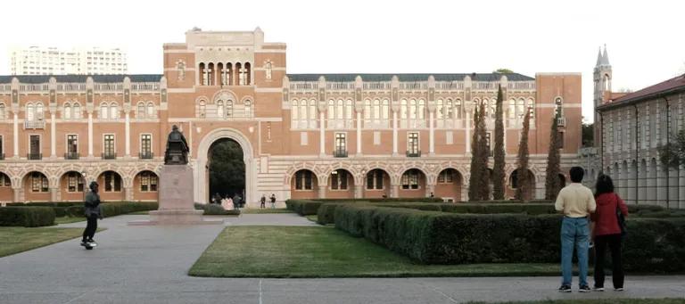 The exterior of Rice University – a large building with greenery and students outside.