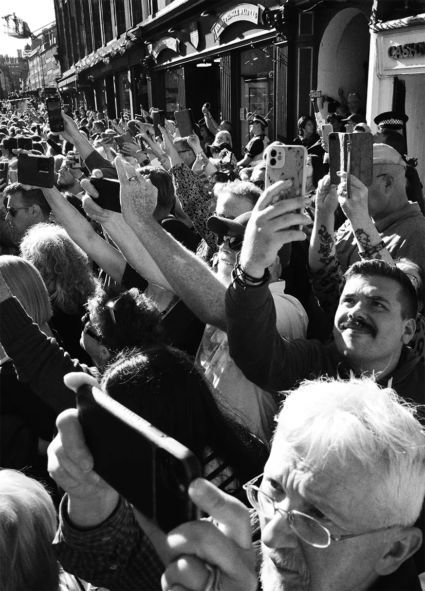 Black and white scene of a crowd of people with their camera phones capturing something out of shot.