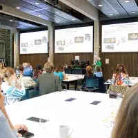 A group of people sitting at tables in a classroom looking up at projector screens.