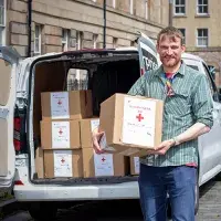 A person holding a box that reads "Humanitarian Aid". A van is in the background with more boxes sitting inside.