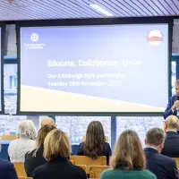 A person in a suit speaking in front of a small group of people. A projector reads "Educate, Collaborate, Unite".