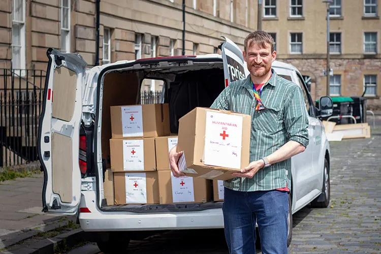 A person holding a box that reads "Humanitarian Aid". A van is in the background with more boxes sitting inside.