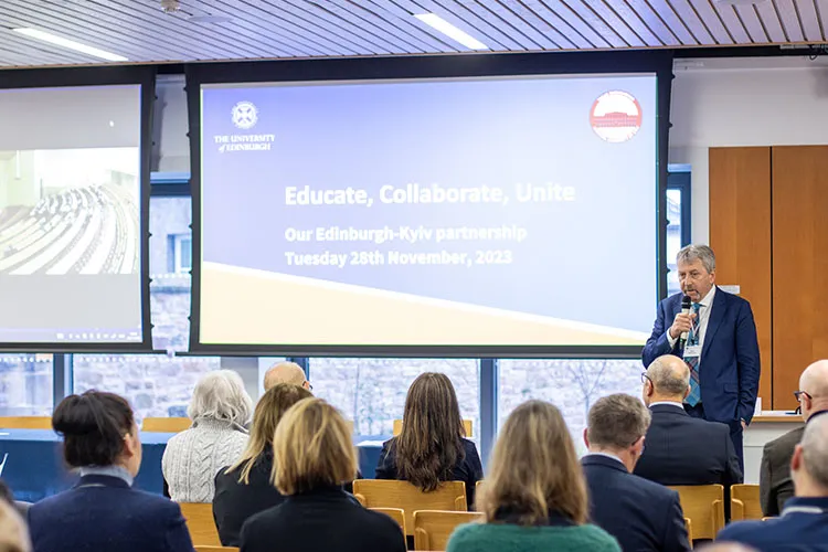 A person in a suit speaking in front of a small group of people. A projector reads "Educate, Collaborate, Unite".