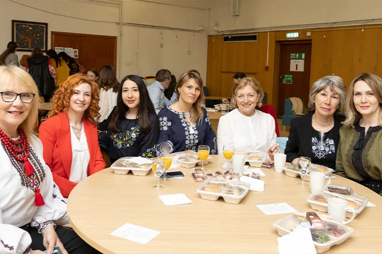 Seven people sitting at a table with food and drinks in the University of Edinburgh's Chaplaincy. Some people wear traditional Ukranian blouses.