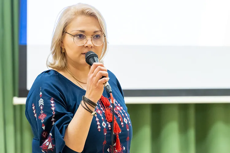 A person wearing a traditional Ukranian blouse speaking with a microphone. A projector screen is visible behind them.