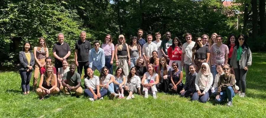 A large group of doctoral students standing outside, smiling at the camera surrounded by green grass and trees.