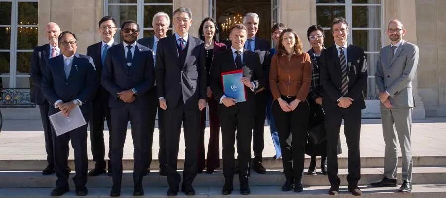 A group of officials in formal business attire standing in a group. At the centre is French President, Emmanuel Macron, holding a binder that reads "U7+".