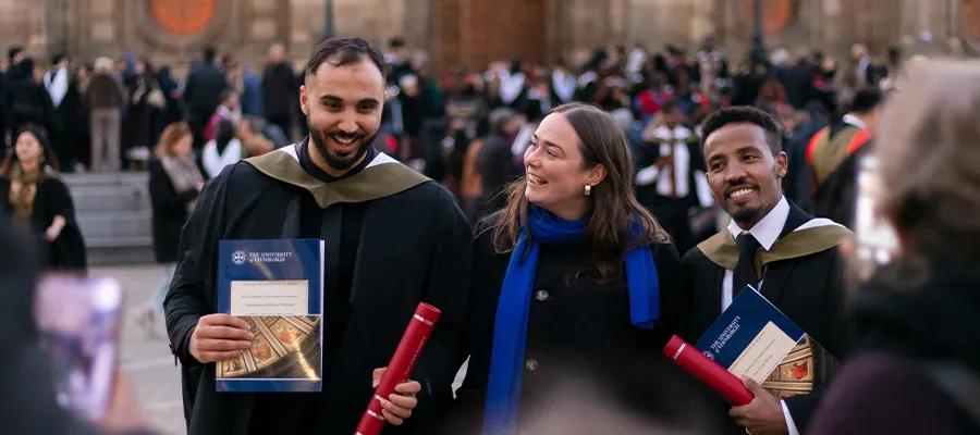 Three people walking and talking in front of a group of graduates. Two of them are holding University branded folders and wearing graduation robes and hats, and one is wearing business attire and a blue scarf.