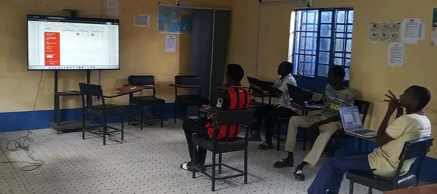 Four people sitting in a room with laptops on their laps, while looking at a large screen.