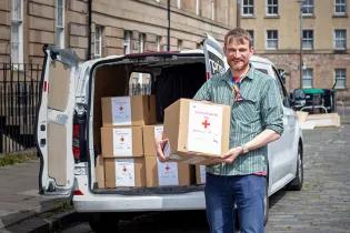 A person holding a box that reads "Humanitarian Aid". A van is in the background with more boxes sitting inside.