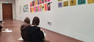 Young people sitting on the ground making art in front of artwork on the wall.