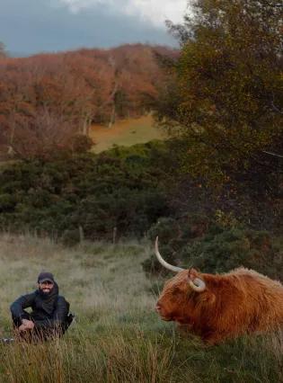 A person sitting in the grass next to a highland cow. Mountains, trees and bushes are visible in the background.