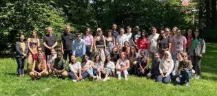 A large group of doctoral students standing outside, smiling at the camera surrounded by green grass and trees.