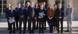 A group of officials in formal business attire standing in a group. At the centre is French President, Emmanuel Macron, holding a binder that reads "U7+".