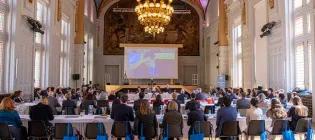 A large group of people sitting at tables in a large, elegant room, looking at information on a screen.